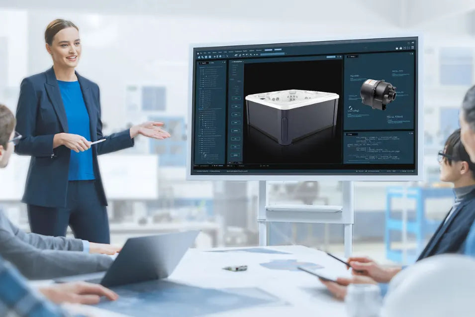 A woman stands at a conference table pointing toward a large screen displaying a 3D rendering of a hot tub product